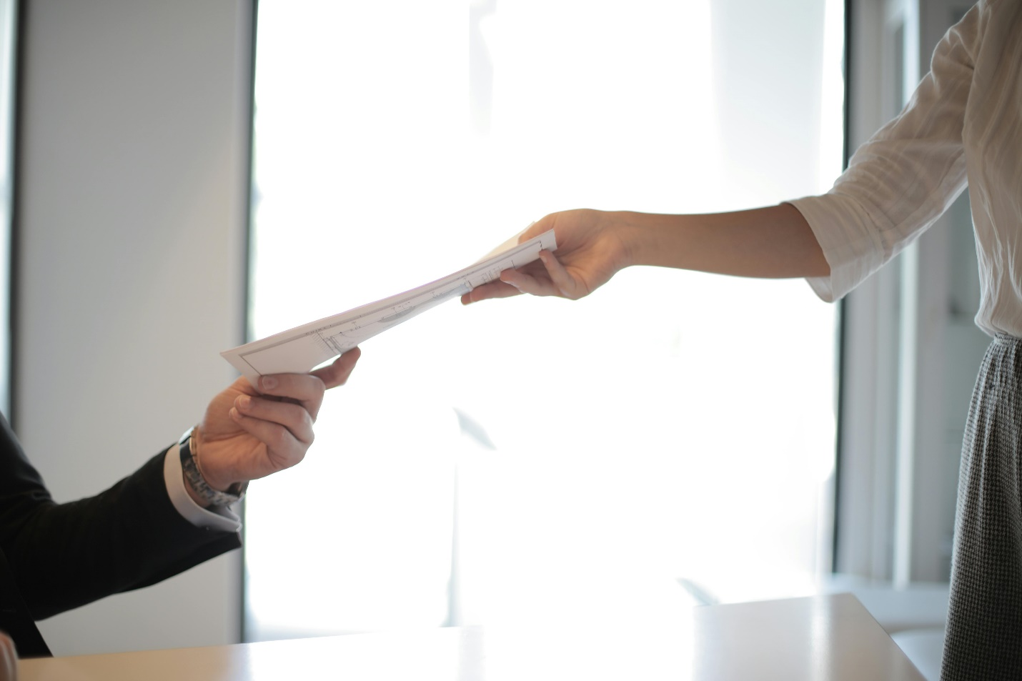 The hand of a person in a suit jacket and watch is seen receiving a paper from another person standing in a white shirt and dark pants by a large window, showing that interviewing techniques are not the only thing required to ascertain a candidate's eligibility for a role.