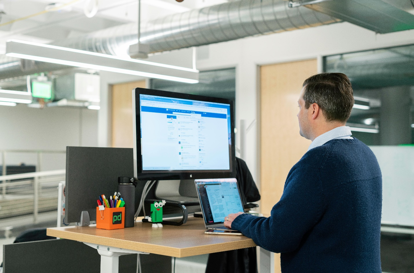 A man in a blue sweater and collared shirt browses a networking platform on a computer monitor while also using a laptop on a small wood and metal desk with stationery, giving a glimpse into how having an online presence can help on-air broadcast talent find recruiters.