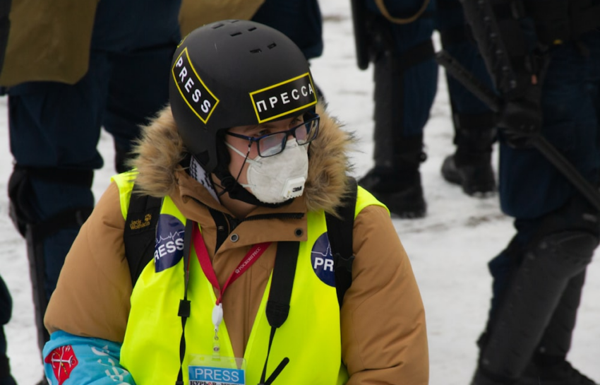 A Journalist Live Reporting While Wearing a Press Vest, Facemask, and Helmet