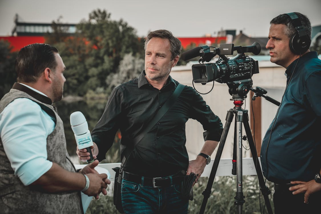 A man holding a microphone while another man stands behind the camera 