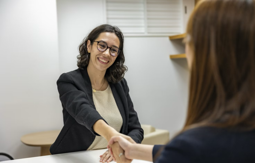 A news channel manager shaking hands with a reporter search expert in a news studio by https://unsplash.com/photos/a-woman-shaking-hands-with-another-woman-sitting-at-a-table-IESB4iFVuzA